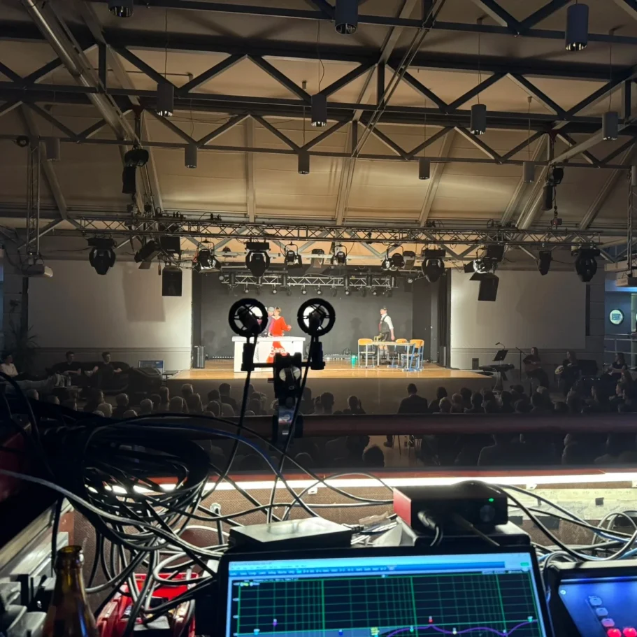 Blick auf eine Bühne mit Publikum, Technik im Vordergrund, beleuchtet von Bühnenlichtern.