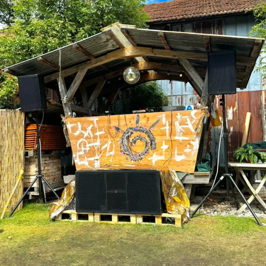 Ein Holzunterstand mit Lautsprechern, einer DJ-Konsole und Pflanzen im Hintergrund.
